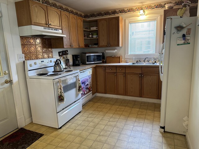 kitchen with brown cabinetry, a sink, light floors, under cabinet range hood, and white appliances