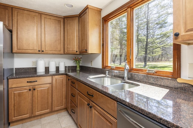 kitchen with stainless steel dishwasher, brown cabinetry, light tile patterned flooring, a sink, and dark stone countertops