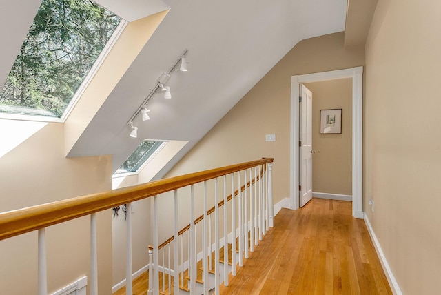 hallway featuring vaulted ceiling with skylight, rail lighting, light wood-style flooring, baseboards, and an upstairs landing