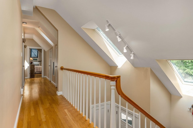 hall with vaulted ceiling with skylight, rail lighting, baseboards, an upstairs landing, and light wood-style floors