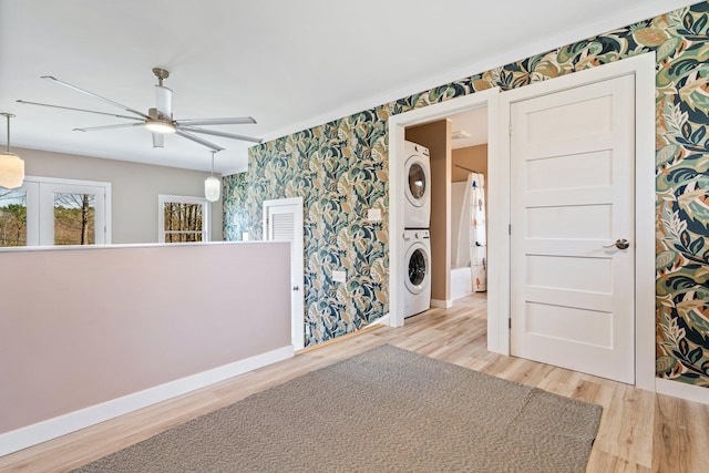 empty room featuring wood finished floors, baseboards, stacked washing maching and dryer, ceiling fan, and wallpapered walls