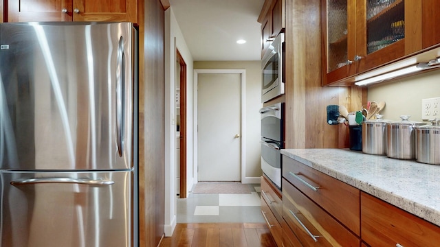 kitchen featuring glass insert cabinets, brown cabinetry, stainless steel appliances, and light stone countertops