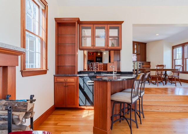 kitchen with a sink, a kitchen breakfast bar, light wood-style flooring, dark stone countertops, and glass insert cabinets