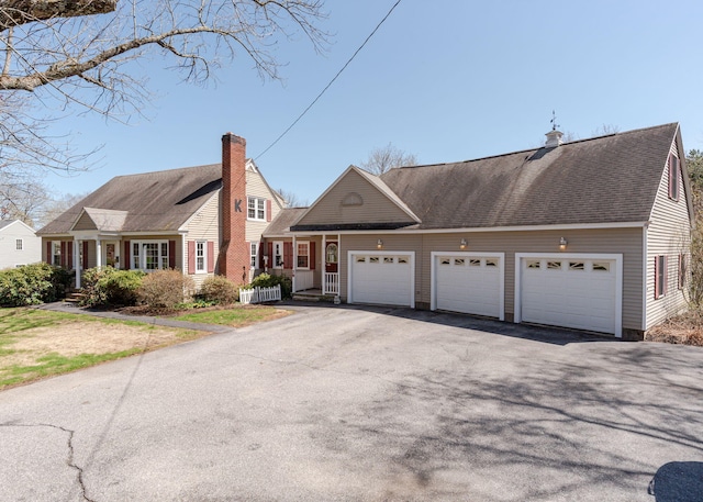 cape cod home featuring a garage, a chimney, roof with shingles, and aphalt driveway