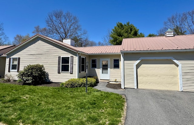 ranch-style home with an attached garage, a front yard, a chimney, driveway, and metal roof