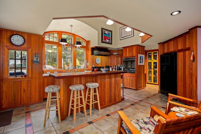 kitchen featuring brown cabinetry, a notable chandelier, a kitchen breakfast bar, black appliances, and exhaust hood