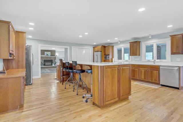 kitchen featuring a brick fireplace, brown cabinets, light wood-type flooring, light countertops, and stainless steel appliances