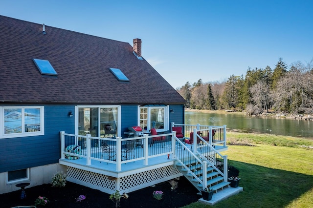 rear view of property with a shingled roof, a deck with water view, a yard, and a chimney