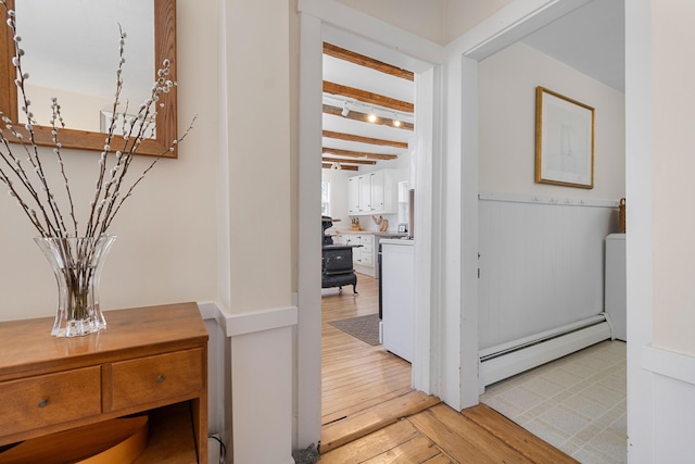 hall featuring light wood-style flooring, beam ceiling, baseboard heating, and wainscoting