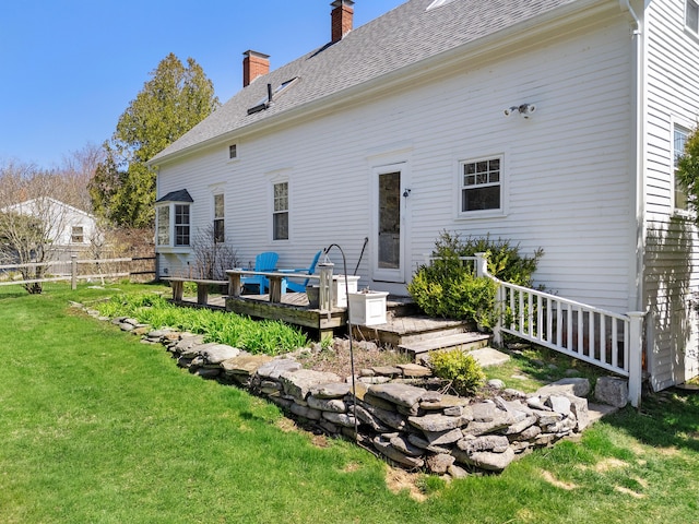 back of property with fence, a shingled roof, a wooden deck, a chimney, and a yard