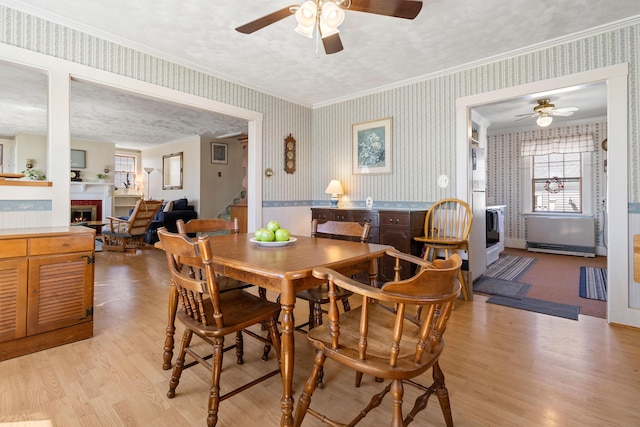 dining space featuring light wood finished floors, a fireplace, a ceiling fan, wallpapered walls, and ornamental molding
