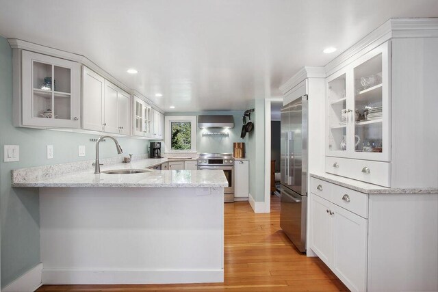 kitchen featuring a peninsula, wall chimney range hood, a sink, light wood-style flooring, and appliances with stainless steel finishes