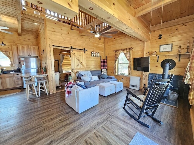 living area with wooden walls, ceiling fan, beamed ceiling, and a wood stove