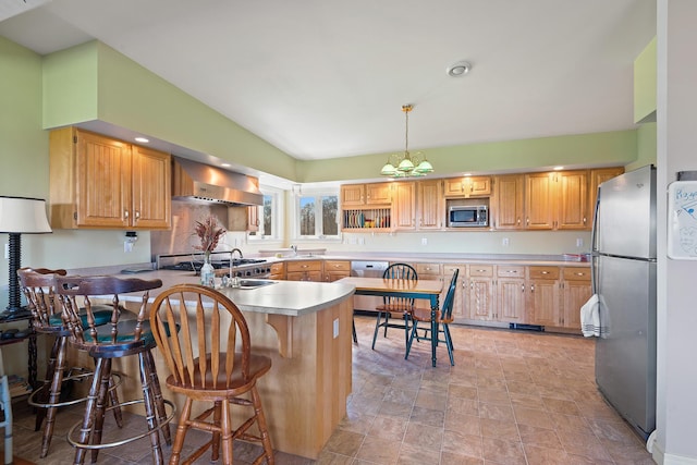 kitchen with a peninsula, a kitchen breakfast bar, wall chimney range hood, a sink, and appliances with stainless steel finishes