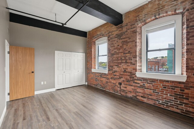 unfurnished bedroom featuring brick wall, wood finished floors, a closet, baseboards, and beamed ceiling