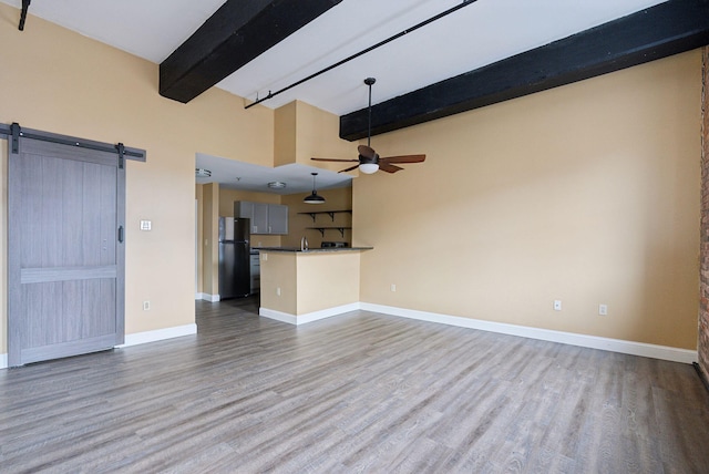 unfurnished living room with a barn door, beam ceiling, ceiling fan, wood finished floors, and baseboards