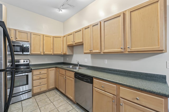 kitchen featuring appliances with stainless steel finishes, a sink, track lighting, dark countertops, and light brown cabinetry