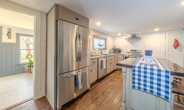 kitchen with appliances with stainless steel finishes, wall chimney exhaust hood, a sink, dark wood-style flooring, and light countertops