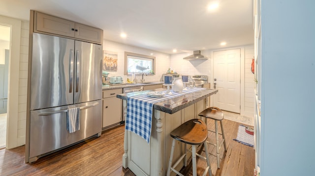 kitchen with appliances with stainless steel finishes, wall chimney range hood, dark wood finished floors, a sink, and a center island