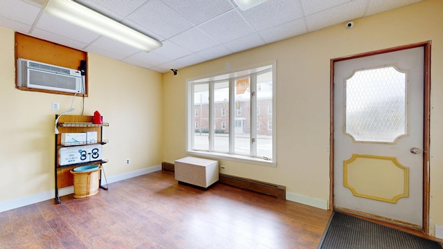 foyer with a baseboard heating unit, wood finished floors, a drop ceiling, a wall mounted air conditioner, and baseboards