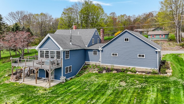 rear view of house with a chimney, a patio, a wooden deck, a lawn, and roof with shingles