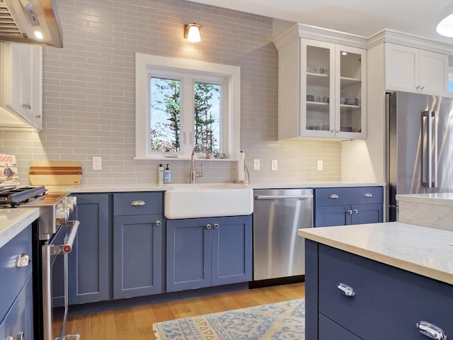 kitchen with appliances with stainless steel finishes, wall chimney exhaust hood, blue cabinetry, a sink, and backsplash