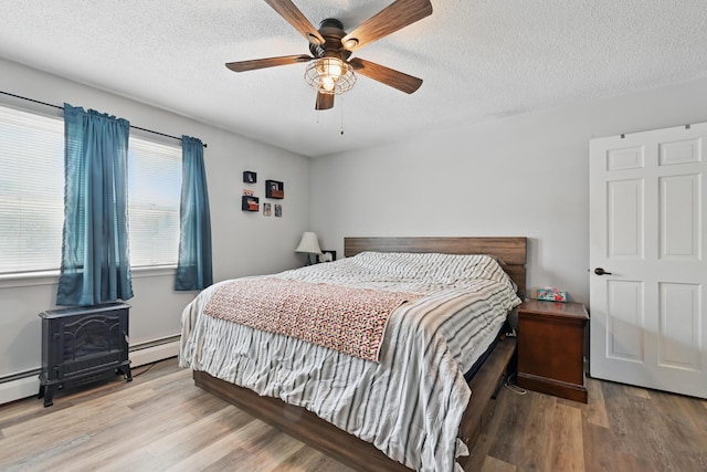 bedroom featuring a wood stove, a textured ceiling, wood finished floors, a baseboard heating unit, and a ceiling fan
