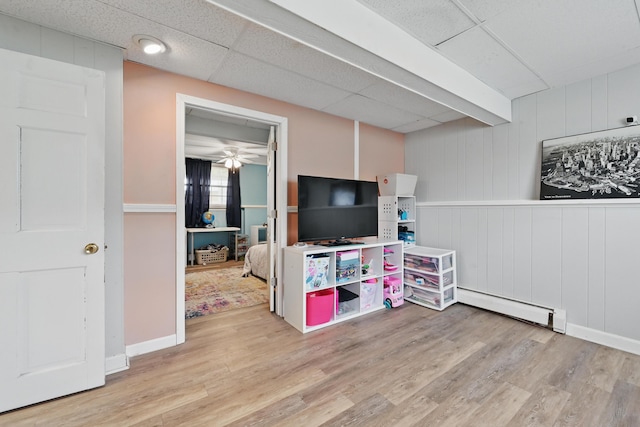 rec room with a baseboard radiator, wood finished floors, a drop ceiling, a ceiling fan, and baseboards