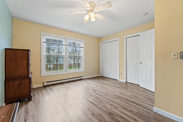 unfurnished bedroom with a baseboard heating unit, multiple closets, baseboards, wood finished floors, and ceiling fan
