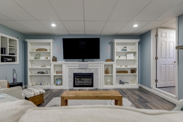 living room with wood finished floors, a drop ceiling, a stone fireplace, baseboards, and recessed lighting