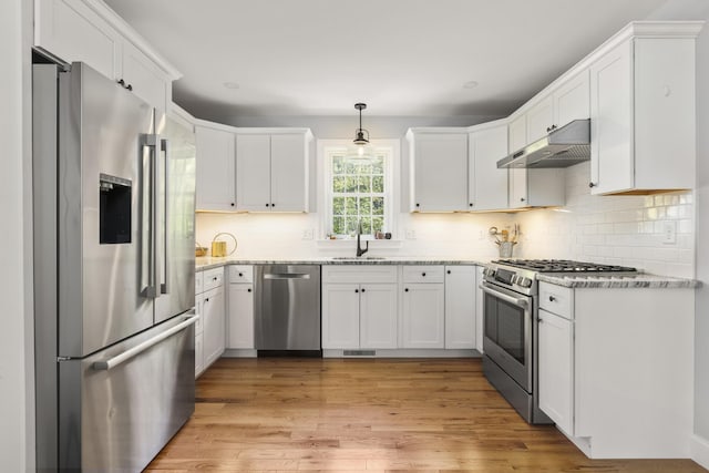 kitchen with stainless steel appliances, under cabinet range hood, a sink, light stone counters, and white cabinets