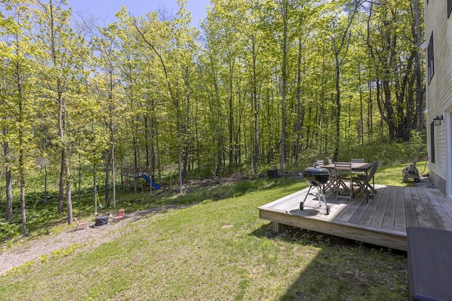 view of grassy yard featuring outdoor dining space, a playground, a wooden deck, and a forest view