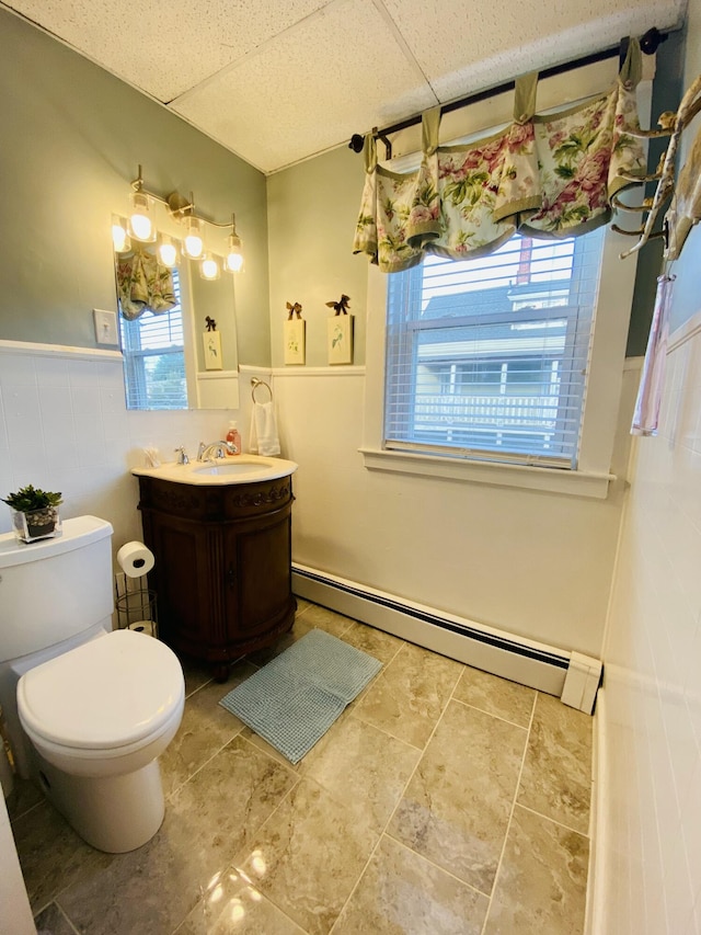 half bathroom featuring baseboard heating, wainscoting, vanity, toilet, and a drop ceiling