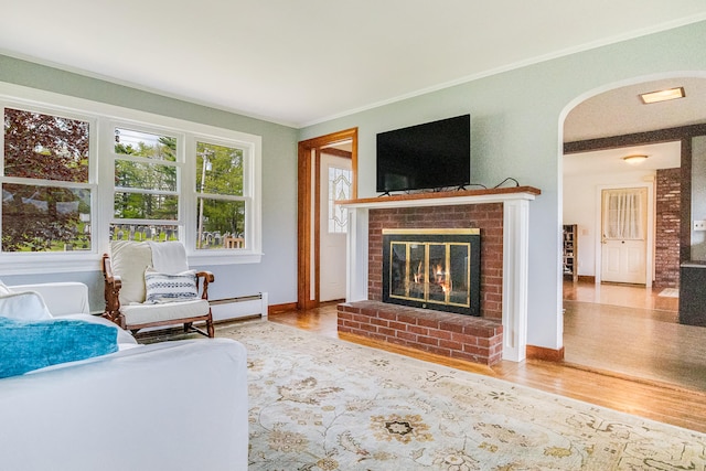 living area featuring a brick fireplace, a baseboard heating unit, wood finished floors, crown molding, and arched walkways