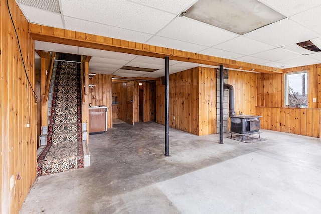 below grade area featuring a paneled ceiling, wooden walls, a wood stove, and stairway