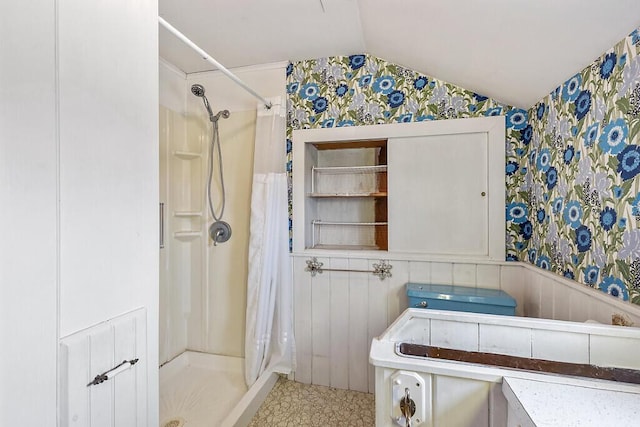 bathroom with wallpapered walls, vaulted ceiling, a shower stall, and wainscoting