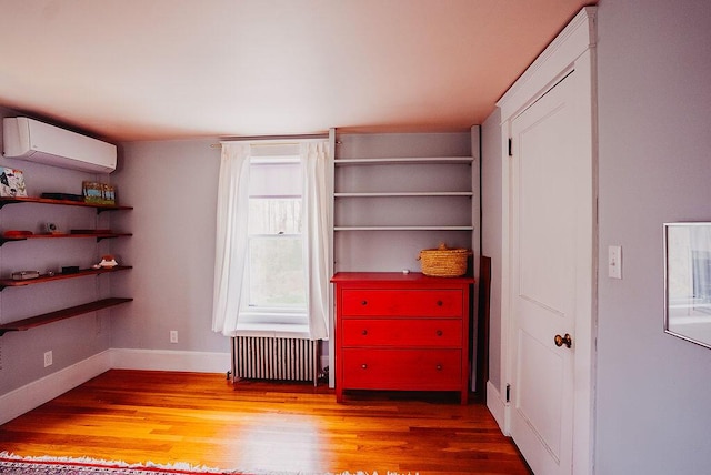 unfurnished bedroom with radiator, a wall unit AC, wood finished floors, and baseboards