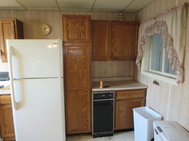 kitchen featuring freestanding refrigerator, wallpapered walls, light countertops, brown cabinetry, and a drop ceiling