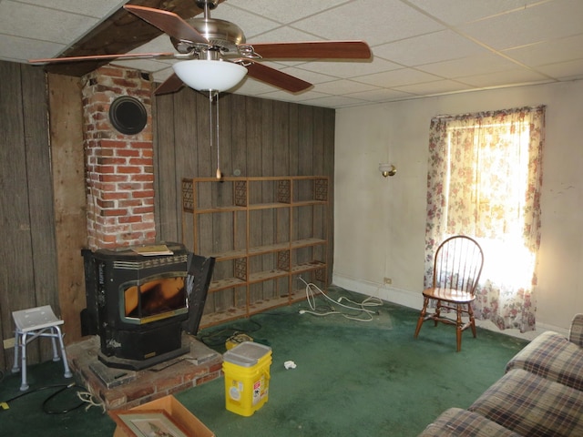 living room with a wood stove, wooden walls, ceiling fan, a paneled ceiling, and carpet
