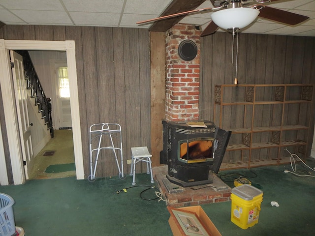 unfurnished living room with a wood stove, wood walls, a paneled ceiling, ceiling fan, and stairs