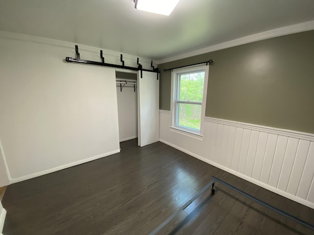 unfurnished bedroom featuring a barn door, dark wood-style flooring, a wainscoted wall, and a closet