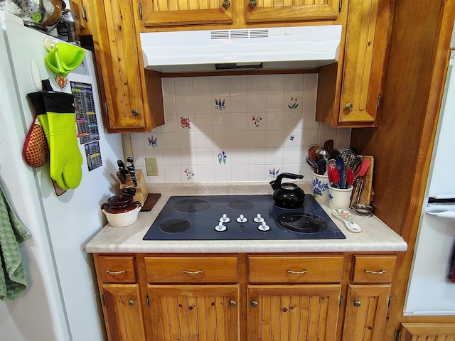 kitchen with freestanding refrigerator, black electric cooktop, under cabinet range hood, tasteful backsplash, and brown cabinetry