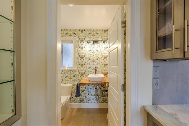 half bathroom with wood finished floors, toilet, vanity, wallpapered walls, and baseboards