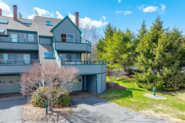 back of house featuring asphalt driveway, a chimney, a balcony, and a shingled roof