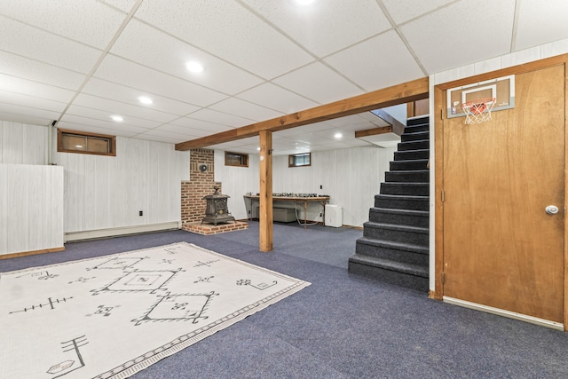 finished basement with a baseboard radiator, stairs, a wood stove, carpet flooring, and a drop ceiling