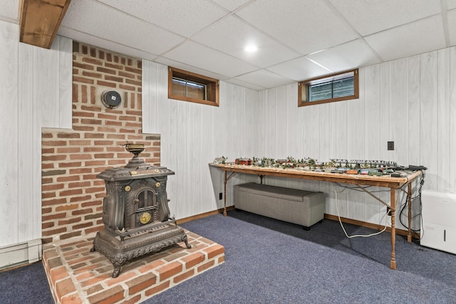 interior space with a wood stove, carpet floors, a baseboard heating unit, a paneled ceiling, and wooden walls