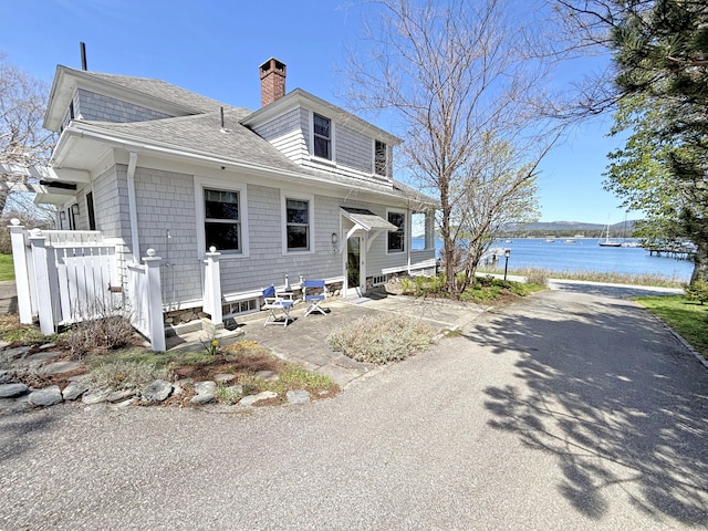 view of front of property with a shingled roof, a chimney, a water view, fence, and a patio