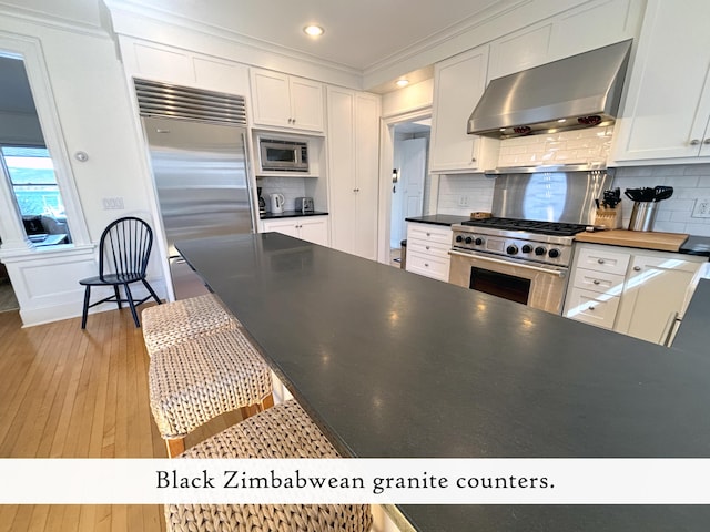 kitchen with wall chimney range hood, built in appliances, light wood-style floors, dark countertops, and white cabinetry