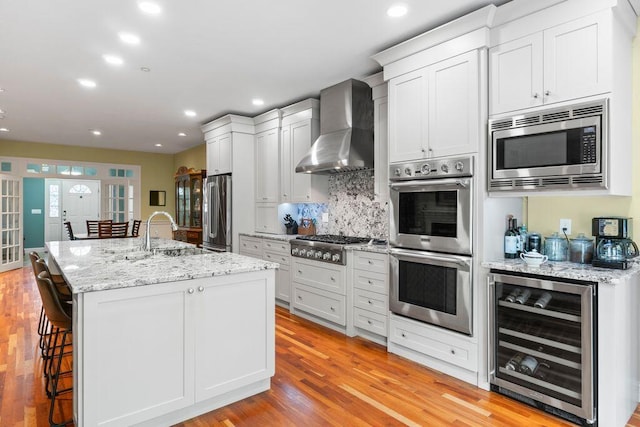 38 Harvest Hill Ln Unit 38, Auburn, ME 04210 - kitchen featuring wine cooler, appliances with stainless steel finishes, wall chimney range hood, light wood-style floors, and a breakfast bar