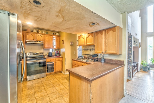 kitchen featuring a peninsula, a sink, under cabinet range hood, a textured ceiling, and appliances with stainless steel finishes